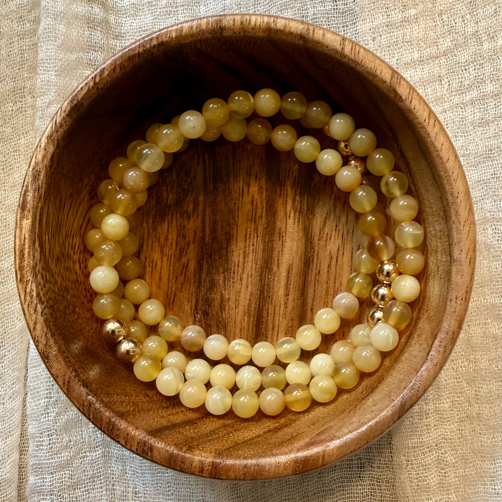 Two beaded stretch bracelets made of pale yellow and cream stone beads with small gold spacer beads, displayed inside a round wooden bowl on a beige fabric surface.
