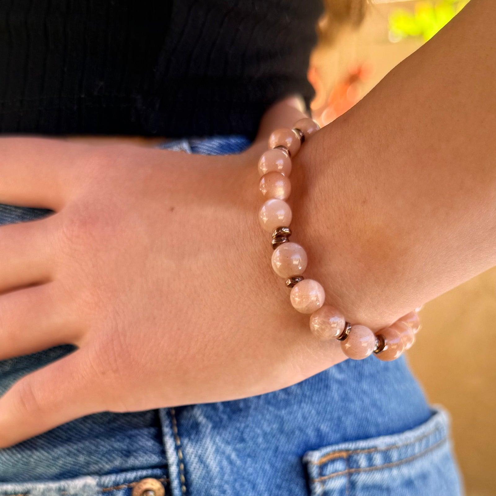 A beaded stretch bracelet made of smooth, round peach-pink stone beads separated by small metallic bronze spacer beads, shown on an outdoor background.