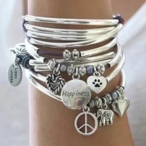A stack of silver charm bracelets worn on a wrist, featuring charms including a peace sign, elephant, paw print, hearts, beads, and a disc engraved with 'Happiness'.