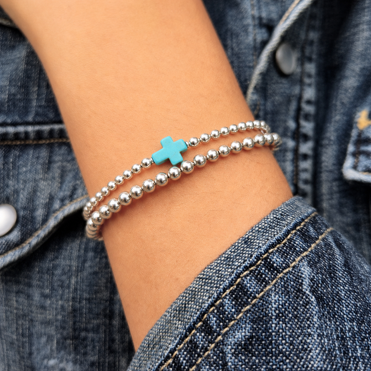 A close-up of a person's wrist wearing two silver beaded bracelets, one with a small turquoise cross charm, styled with a denim jacket.