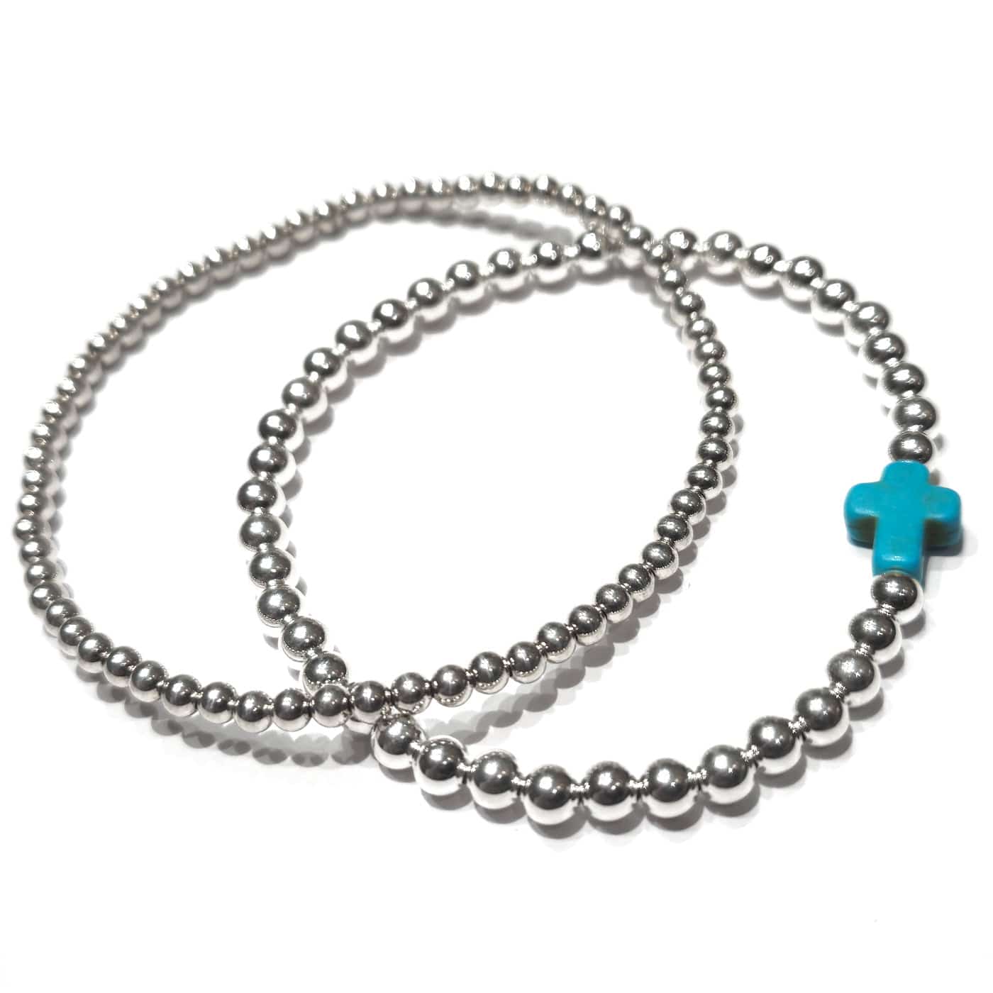 A close-up of a person's wrist wearing two silver beaded bracelets, one with a small turquoise cross charm, styled with a denim jacket.
