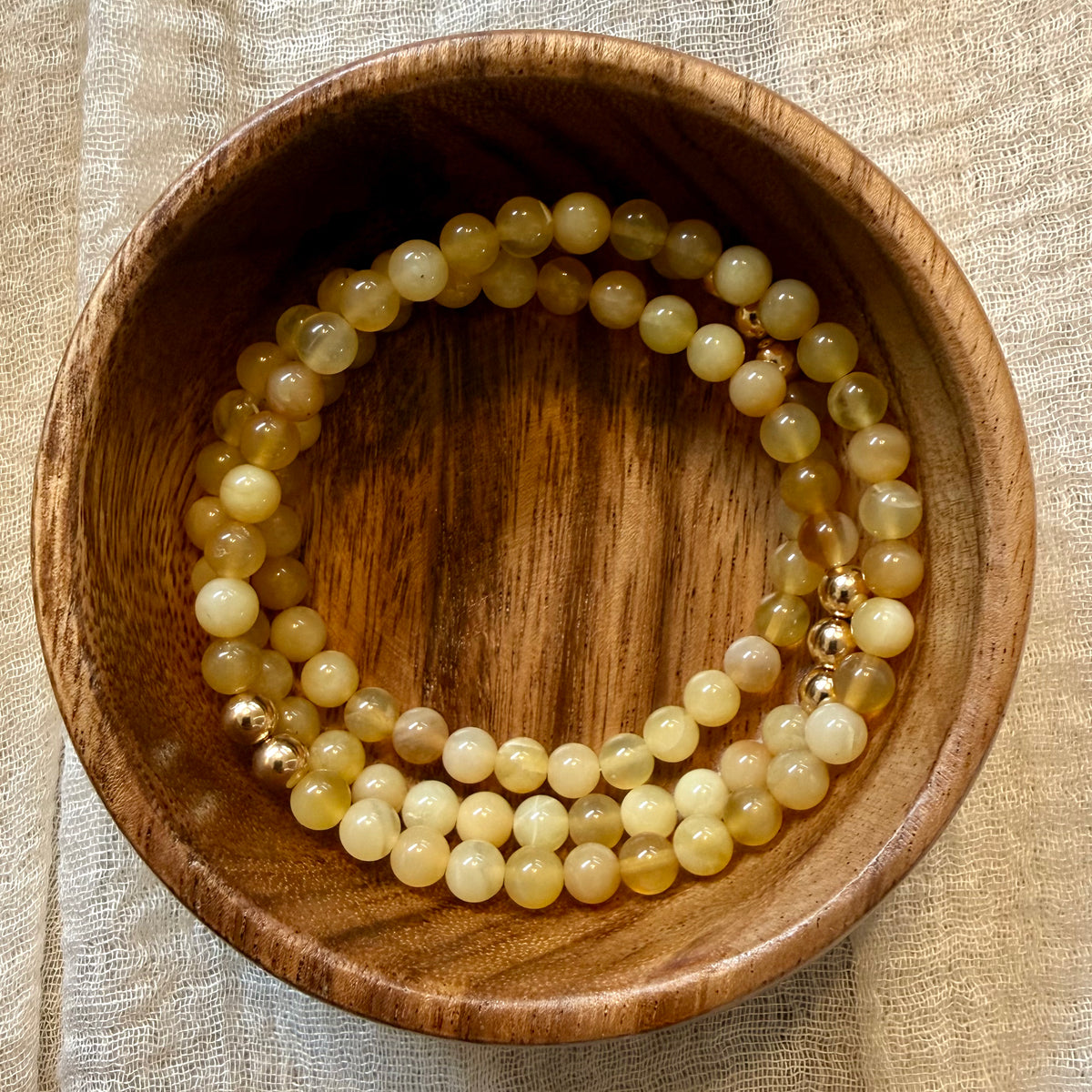 Two beaded stretch bracelets made of pale yellow and cream stone beads with small gold spacer beads, displayed inside a round wooden bowl on a beige fabric surface.