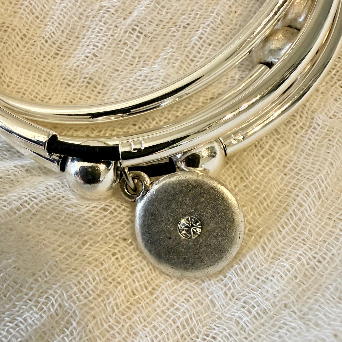 A close-up of a silver-toned bangle bracelet with a round disc charm featuring a small starburst detail, resting on a textured off-white fabric background.