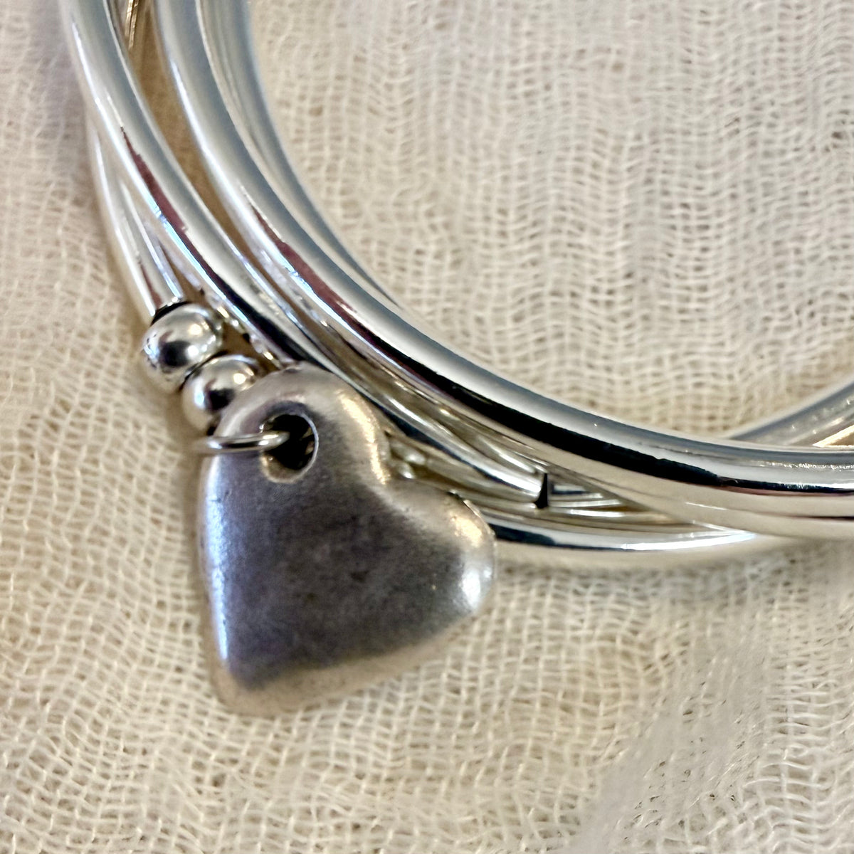 A close-up of a silver multi-strand bangle bracelet with a small hammered heart charm and three silver beads, resting on a textured cream linen fabric.