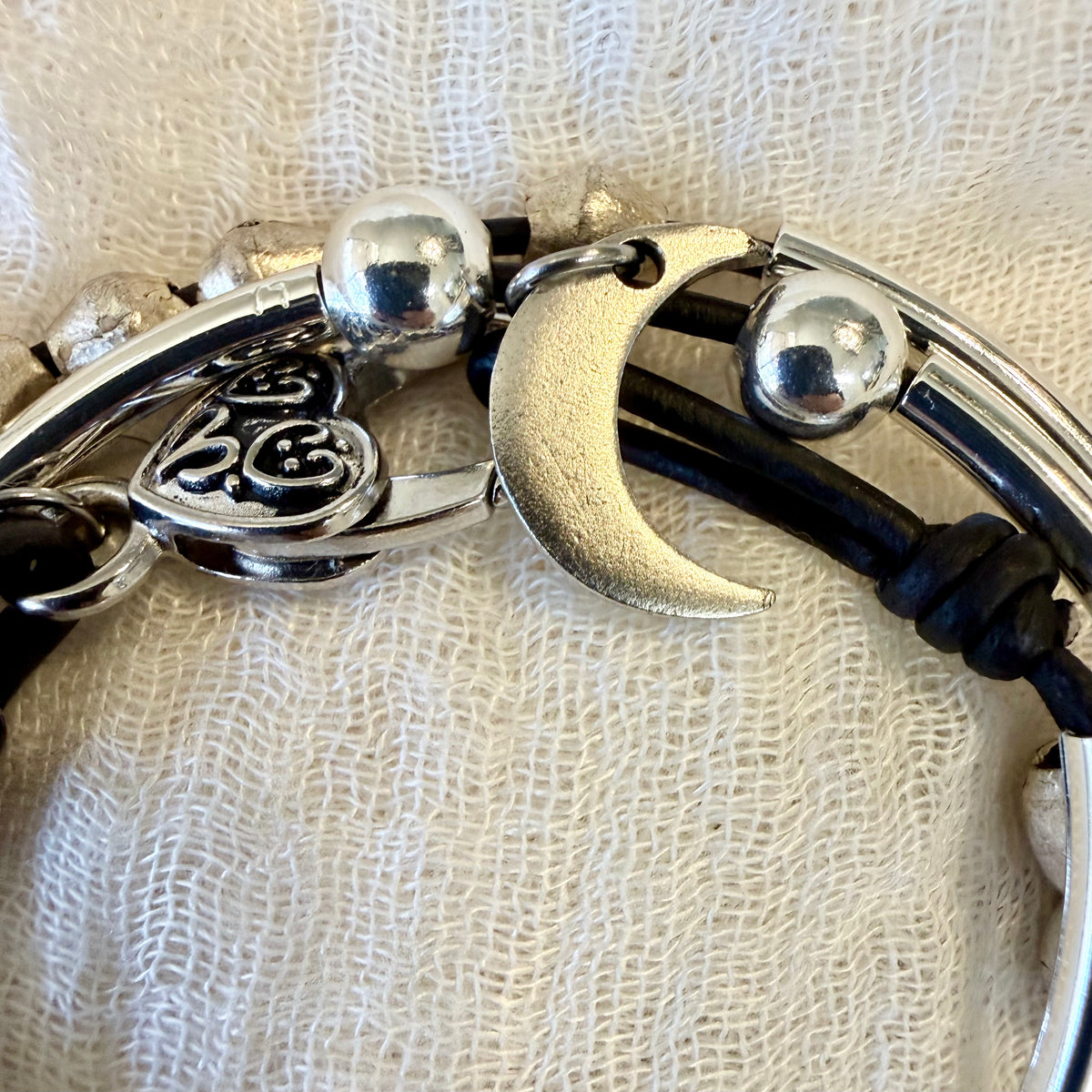 A close-up of a bracelet featuring a silver crescent moon charm, a heart-shaped charm with engraved lettering, polished silver beads, and black cords, displayed on a textured beige fabric background.