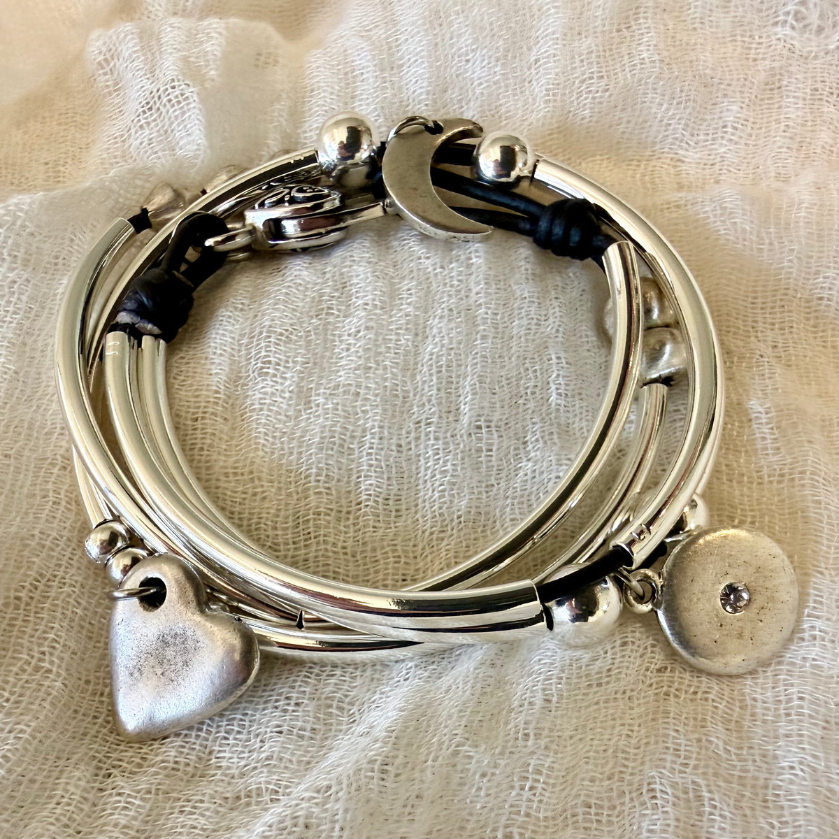 A stack of silver-toned bangles and bracelets with charms, including a heart, crescent moon, and round disc, resting on a textured off-white fabric background.