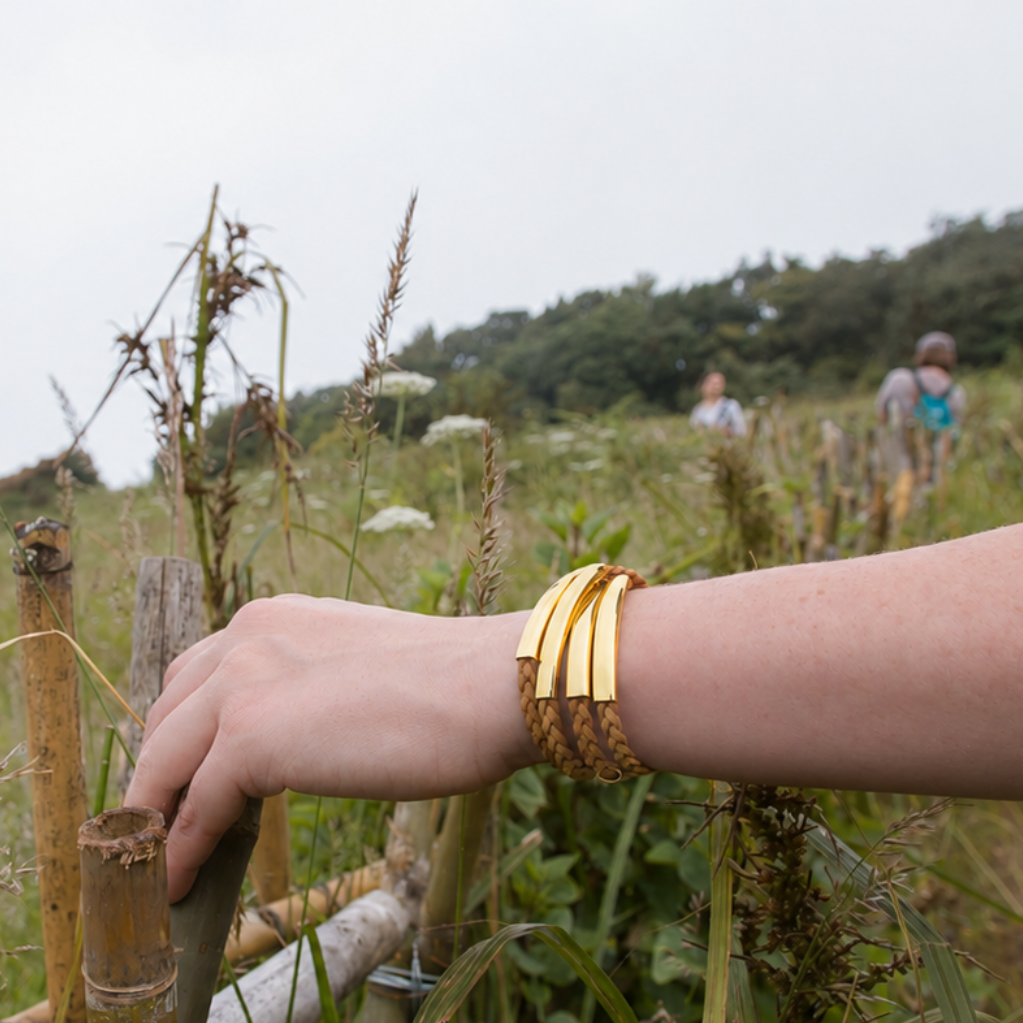 A close-up of a person's hand wearing a stack of wide, polished gold bangles with textured woven trim, set against an outdoor grassy field background.