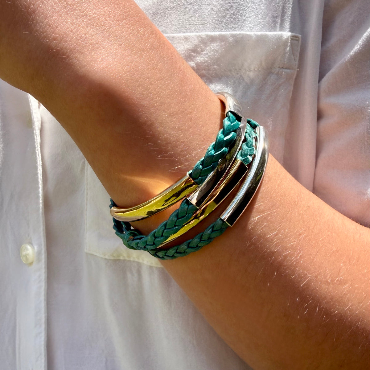 A close-up of a person's wrist wearing a stack of bracelets, including a teal braided wrap bracelet, a polished gold bangle, and a silver-toned bar bracelet, over a white button-down shirt.