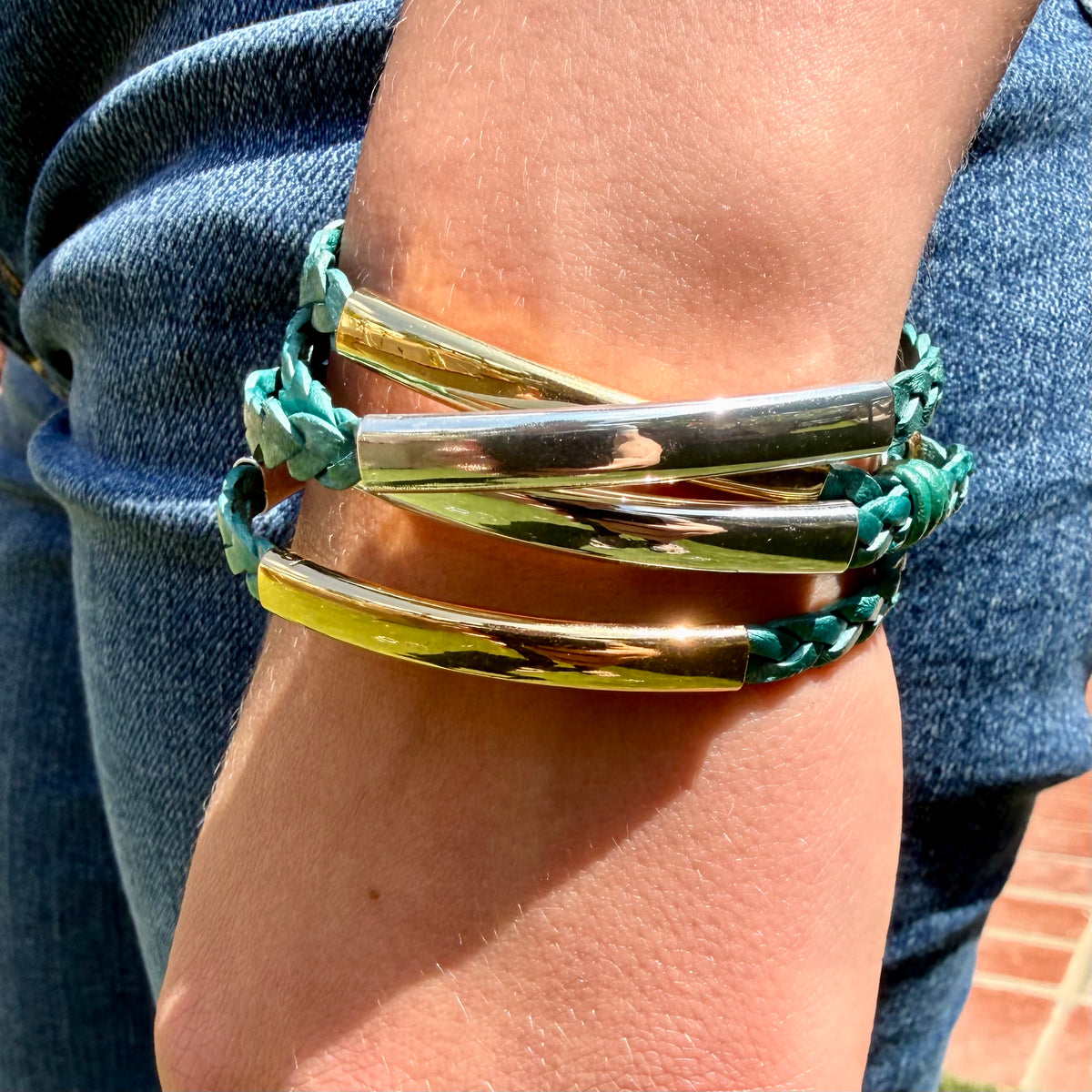 A close-up of a person's wrist wearing a stack of bracelets, including shiny gold-toned metal tubes and teal braided cord bracelets, with blue jeans visible in the background.