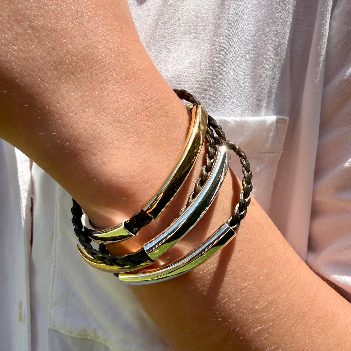 A close-up of a person's wrist wearing a stack of four bracelets: two polished gold-toned bangles, one silver-toned bangle, and one black braided cord bracelet, against a white shirt background.
