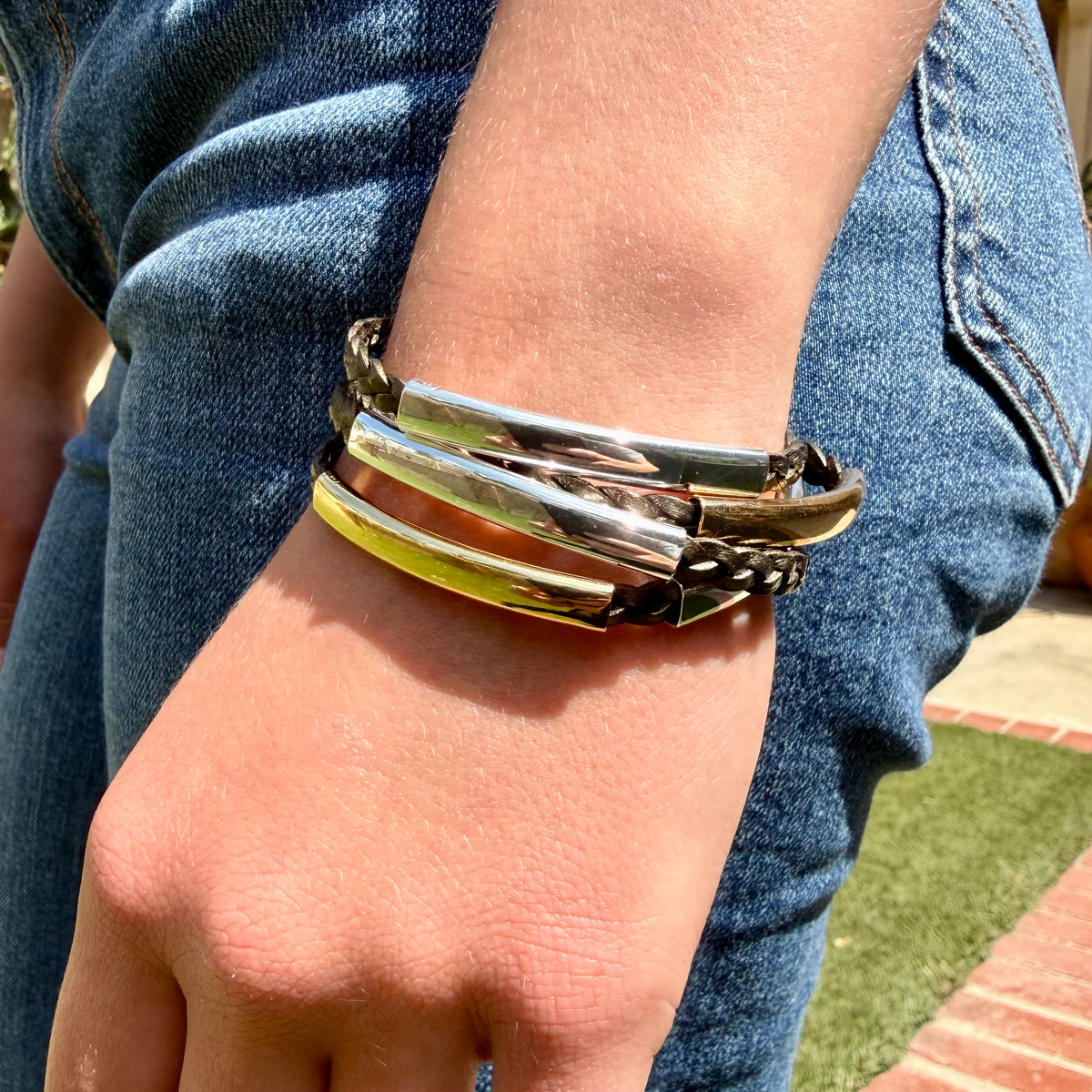 A close-up of a person's wrist wearing a stack of three metallic bangles, including two silver-toned and one gold-toned bangle, paired with blue denim jeans.
