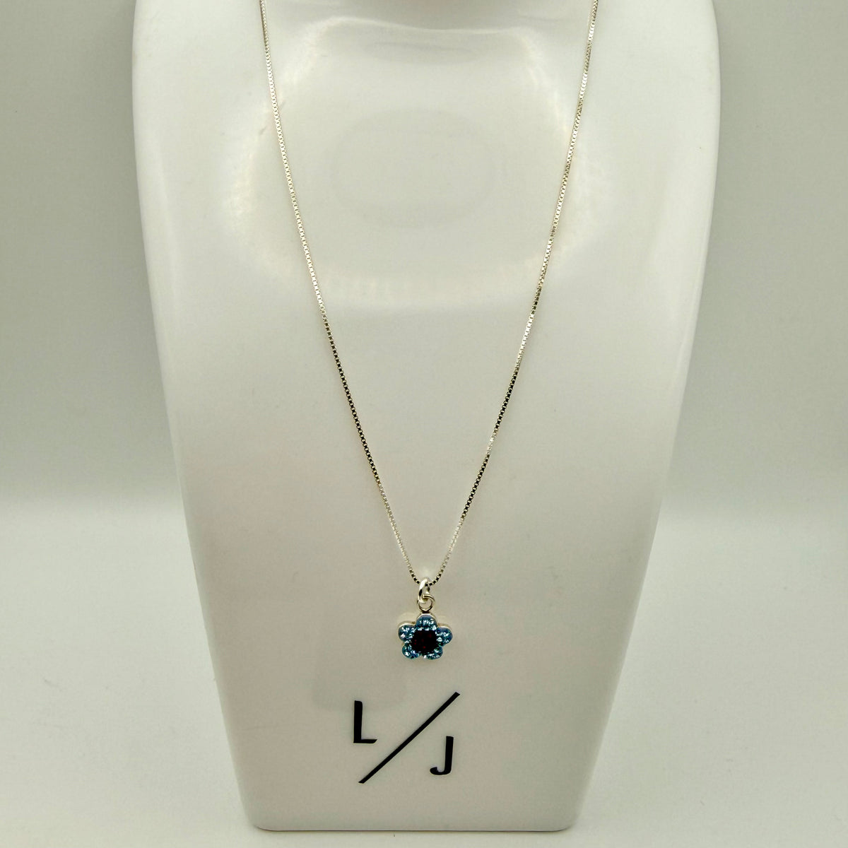 Necklace with a blue pendant on a white stand with 'LJ' initials.