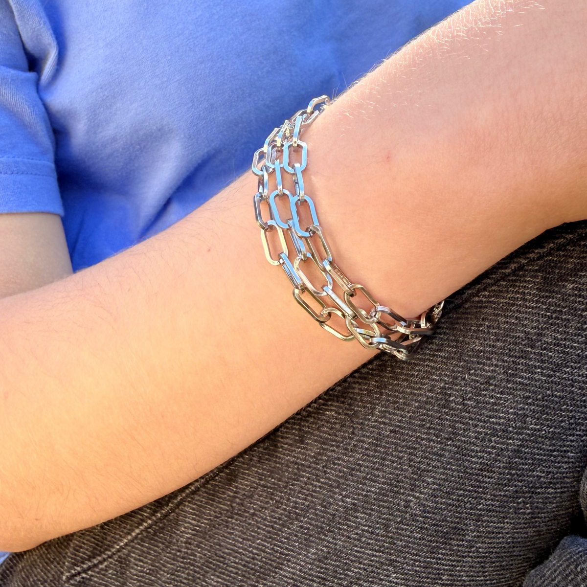 Person wearing a silver chain bracelet on an arm with a blue shirt and gray pants.