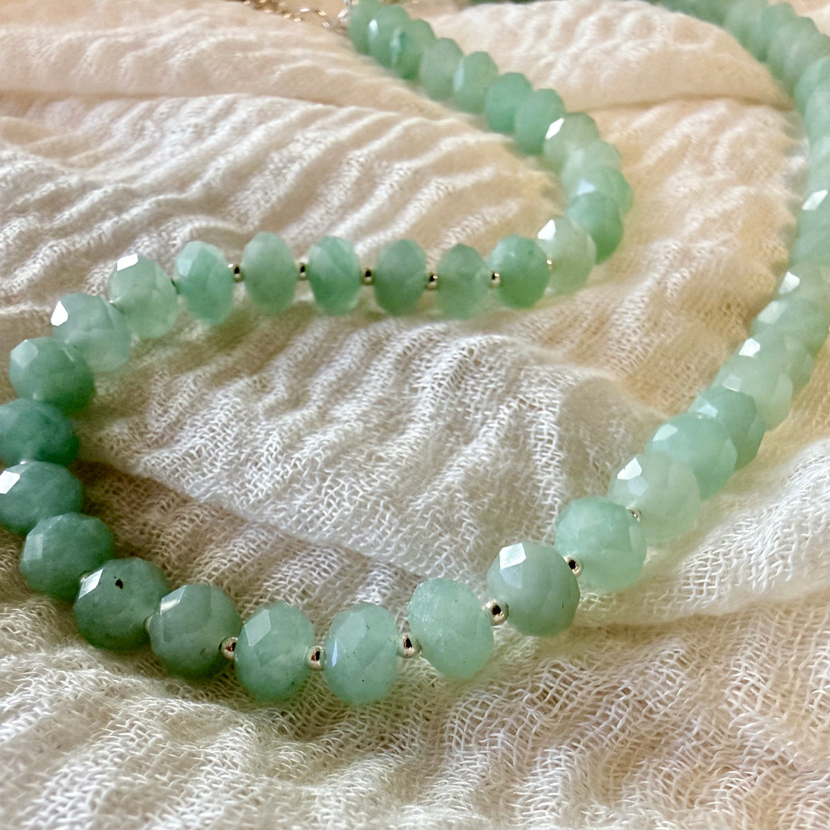 Faceted light green jade bead necklace with small silver spacer beads, draped on a textured cream-colored fabric.