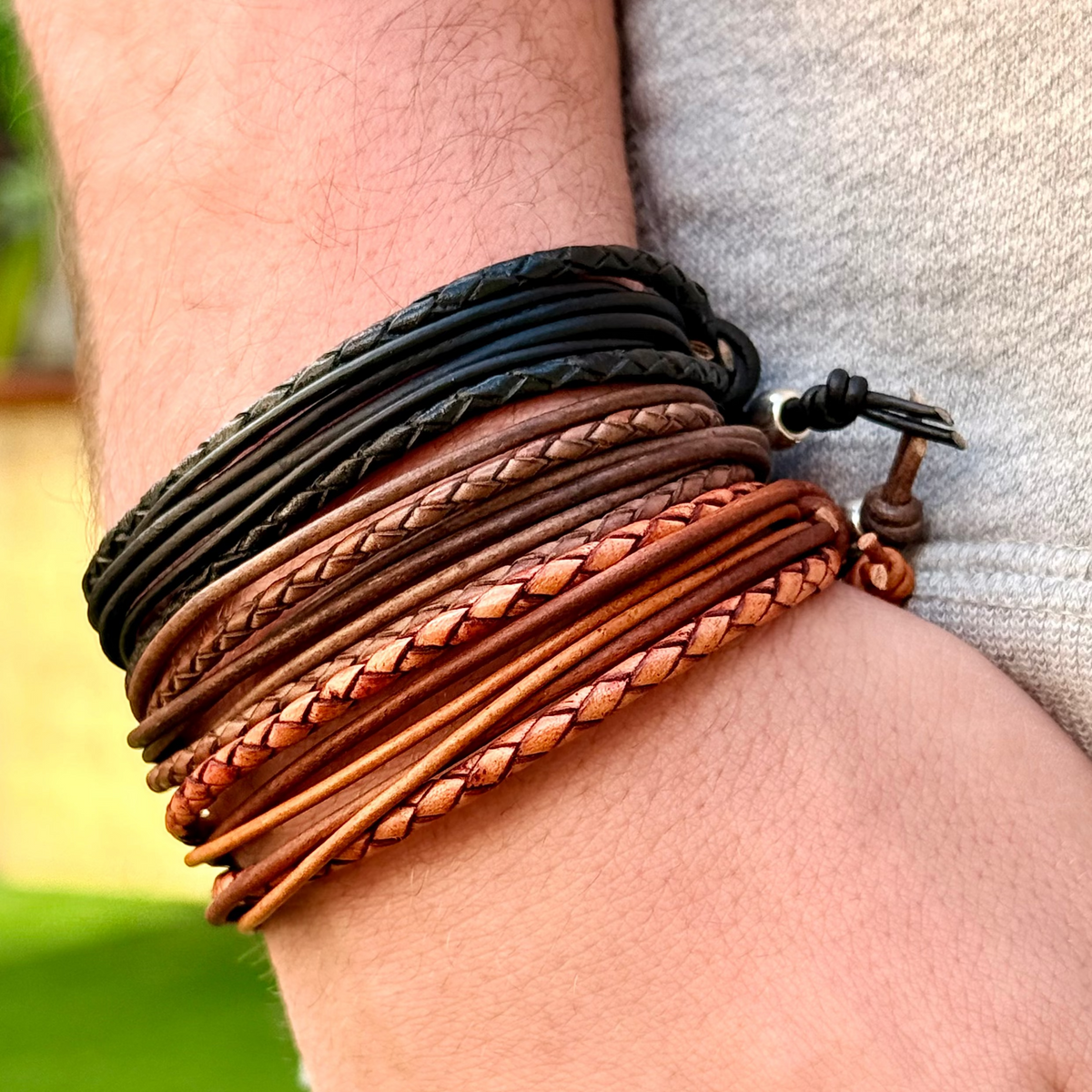 Close-up of a wrist wearing multiple braided leather bracelets with a blurred green background.