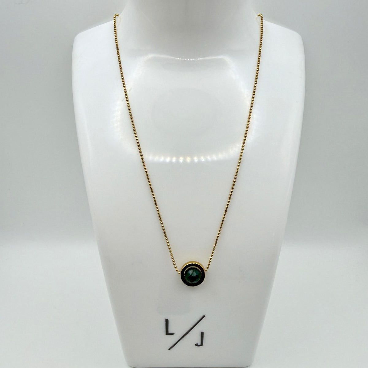 Gold necklace with a green pendant on a white mannequin bust with 'LJ' branding.