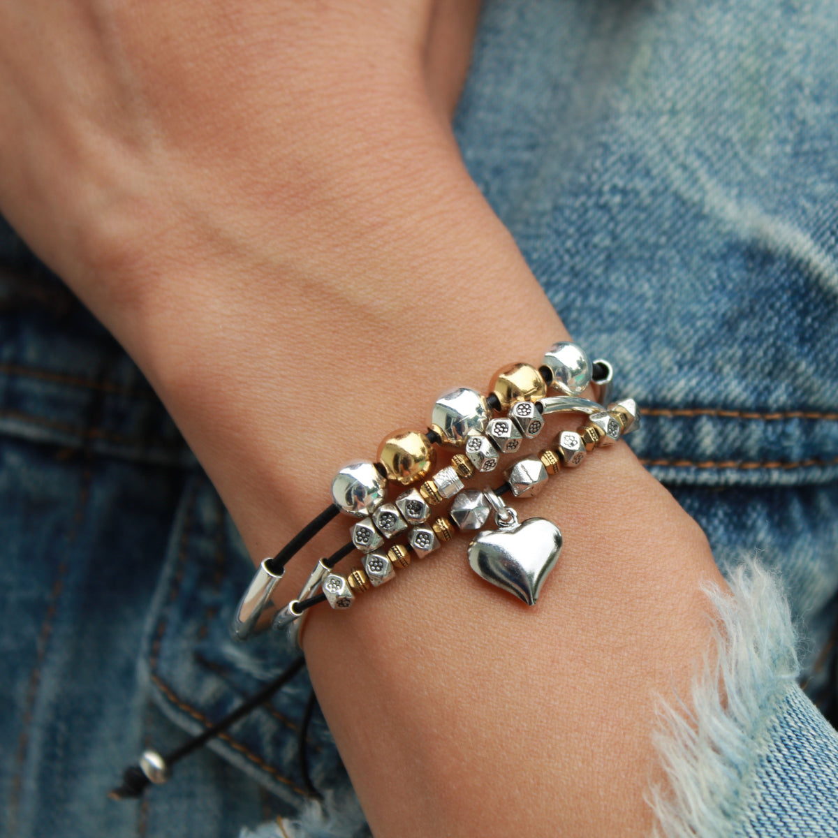 Close-up of a wrist wearing multiple bracelets with heart and geometric designs, against a denim background.