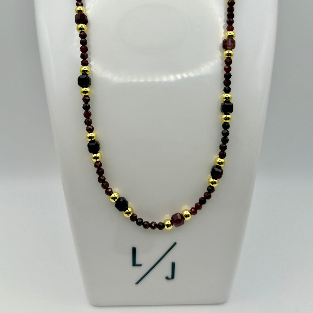 Necklace with red, black, and gold beads on a white stand with 'LJ' branding.