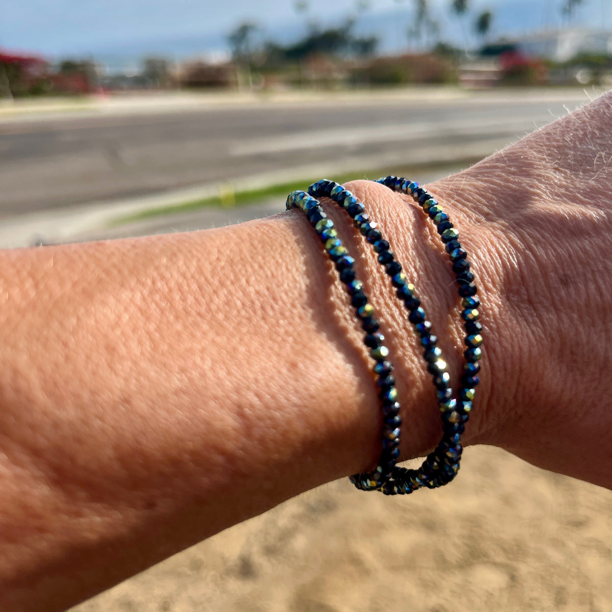 A close-up of a person's wrist wearing three beaded wrap bracelets with iridescent blue and green stones, photographed outdoors with a blurred beach or street background.