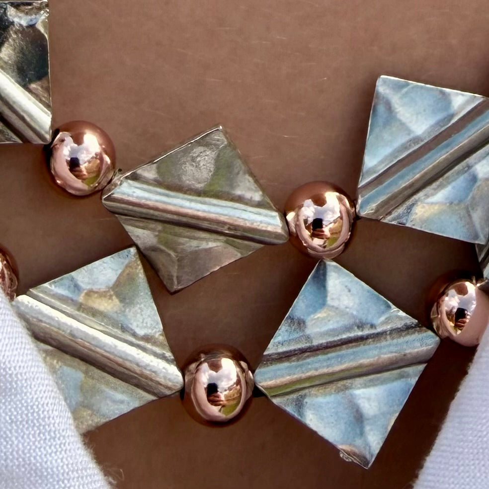 A close-up of a metallic geometric accessory featuring faceted silver triangles and shiny rose-gold spherical accents on a brown background.