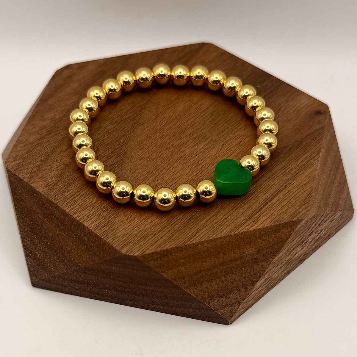 Gold beaded bracelet with a green heart charm on a wooden stand
