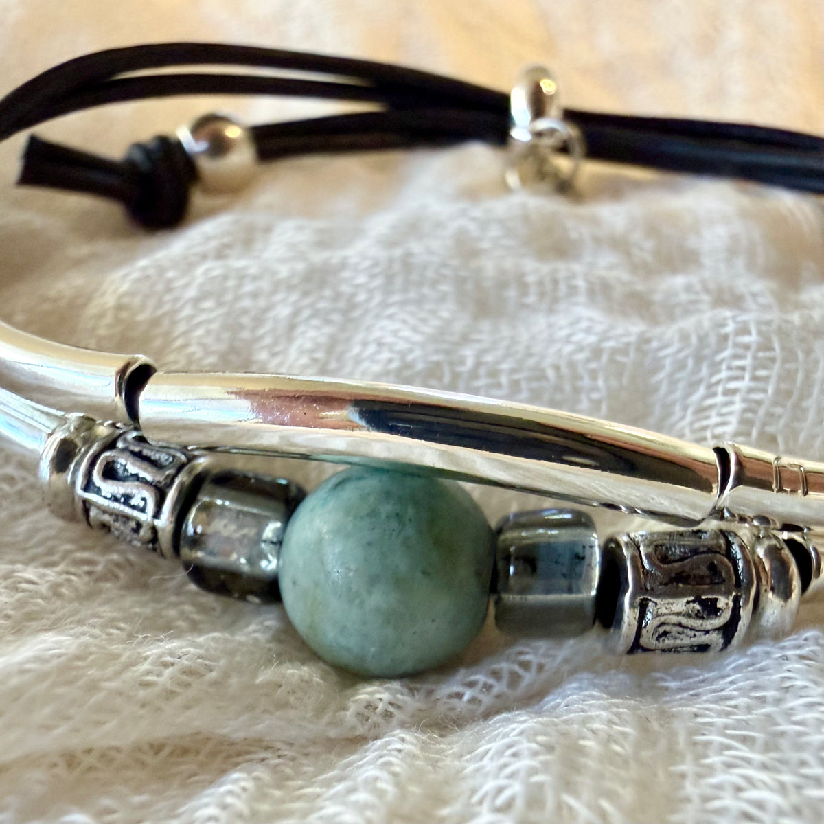 A close-up of a beaded bracelet featuring a central round light blue-green bead, flanked by textured silver spacer beads and a polished silver-toned bar, resting on a white textured fabric background.