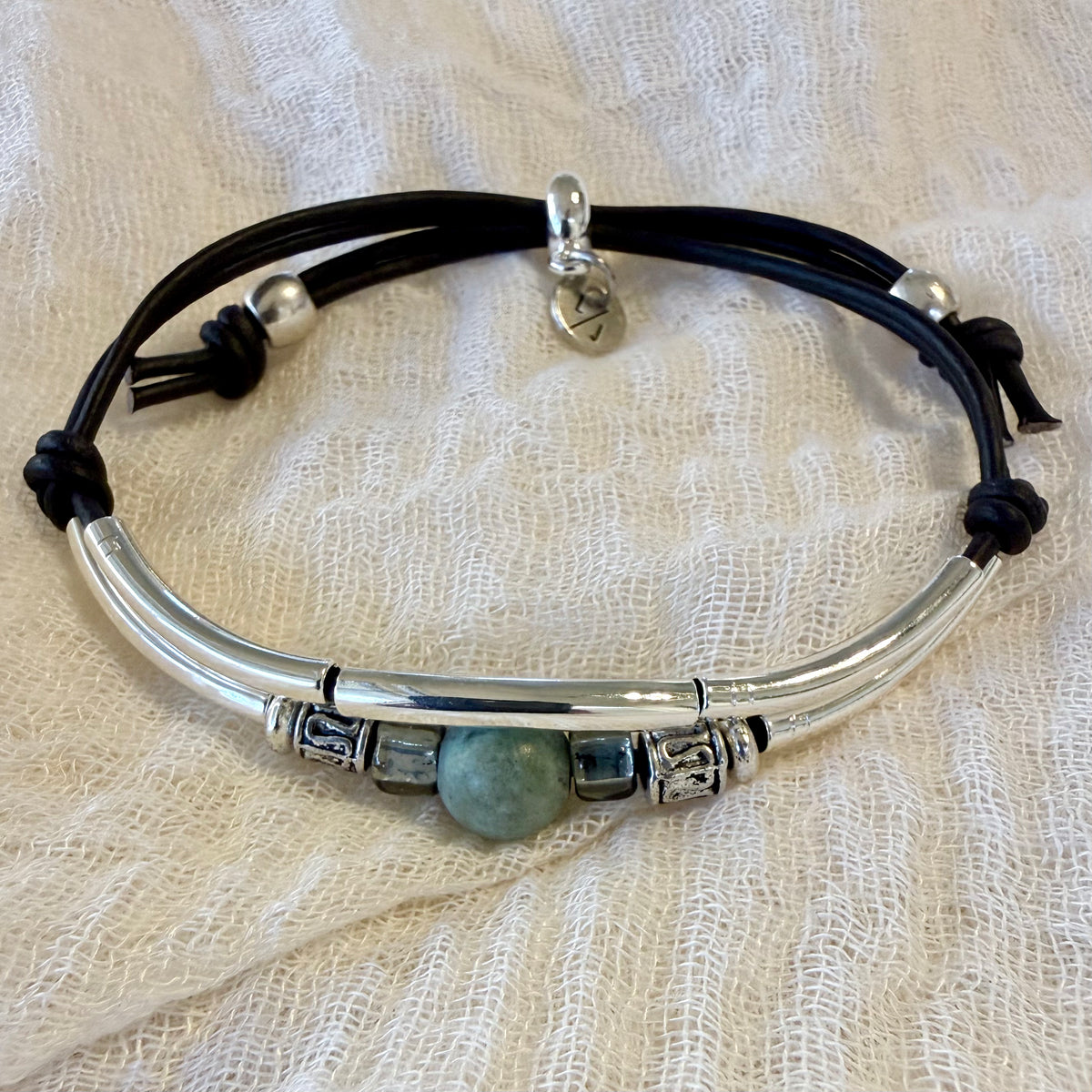 A silver-toned bangle bracelet with a central green stone bead, decorative spacer beads, and black cord wraps, displayed on a textured cream fabric background.