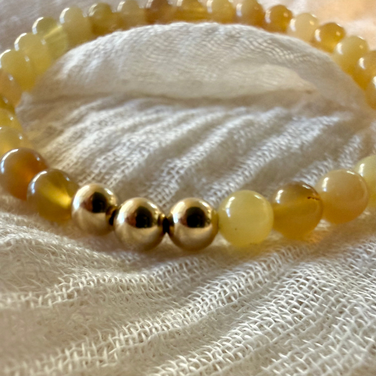 A close-up of a beaded bracelet with translucent pale yellow beads and shiny metallic gold beads, resting on a textured cream fabric background.