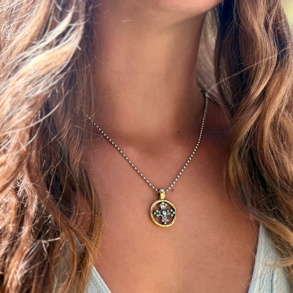 A close-up lifestyle shot of a woman wearing a gold and silver beaded necklace with a circular pendant featuring a floral pattern of green, blue, and white stones.