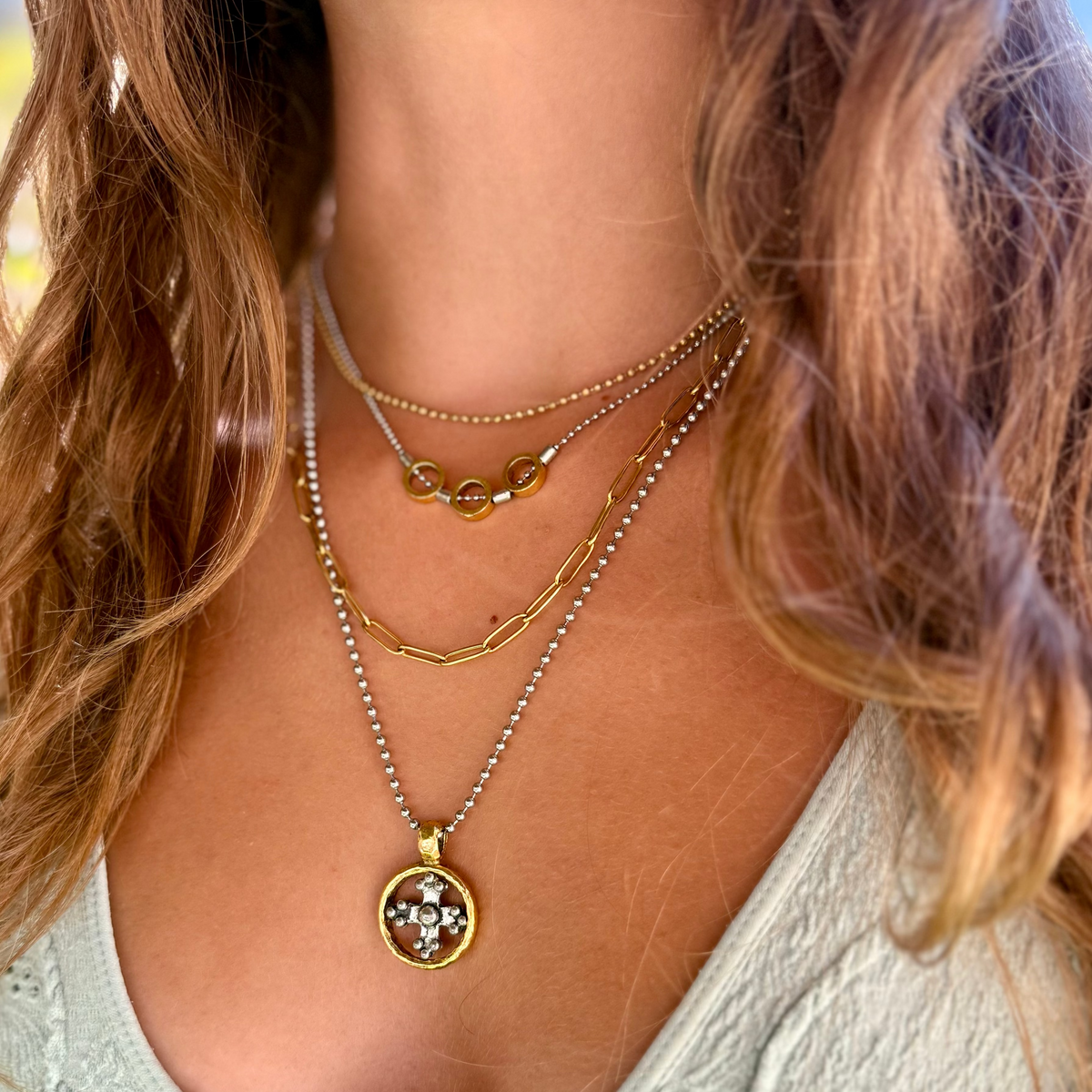 A close-up of a woman wearing a layered necklace set. The set includes a gold chain with a round pendant featuring a white floral design with a small central stone, a silver beaded chain, a gold wavy chain, and a delicate gold chain with small gold rings.