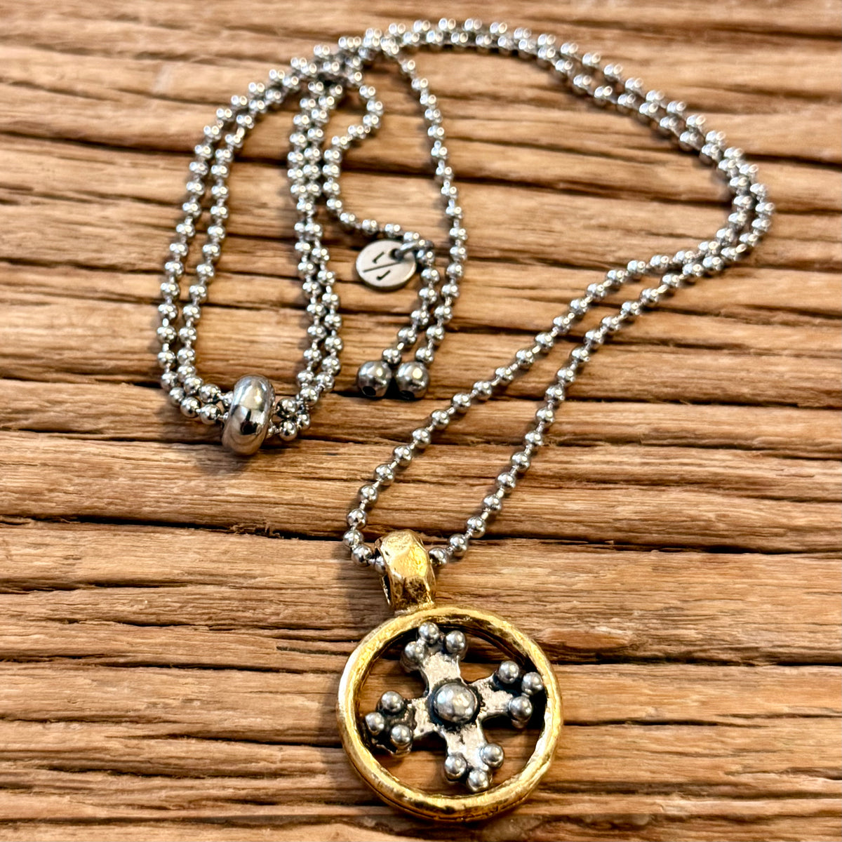A silver ball chain necklace with a round gold-tone pendant featuring a silver cross, displayed on a rustic wooden surface.