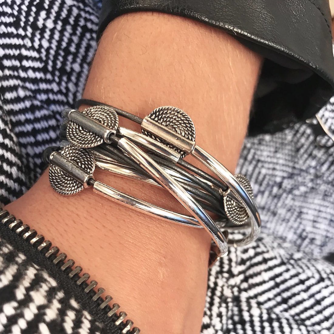 A close-up of a silver wrap bracelet with textured circular details, worn on a person's wrist over a black and white patterned jacket.
