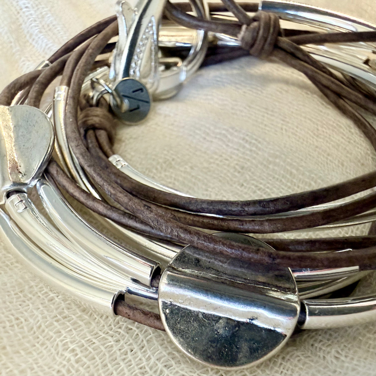 A close-up of a multi-strand bracelet featuring dark brown leather cords and polished silver-tone metal bars, clasps, and a large oval charm, resting on a textured beige fabric background.