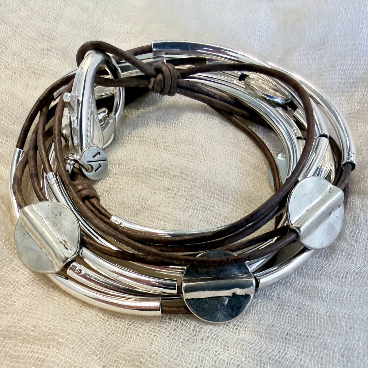 A stack of multi-strand bracelets featuring polished silver-toned metal tubes and dark brown leather cords, displayed on a textured beige fabric background.