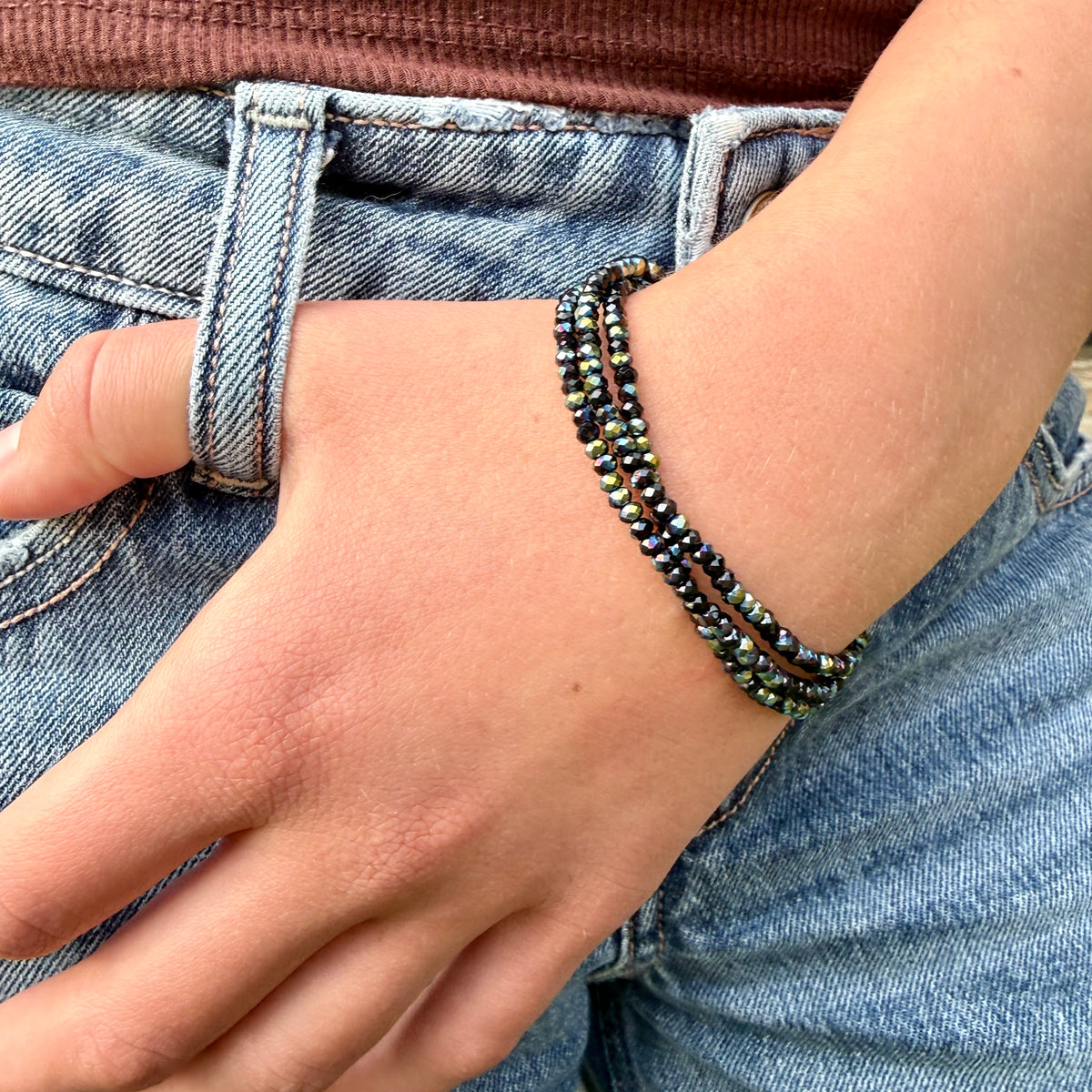 A close-up of a person's wrist wearing a multi-strand beaded bracelet with dark metallic beads and small iridescent green accents, paired with blue denim jeans.