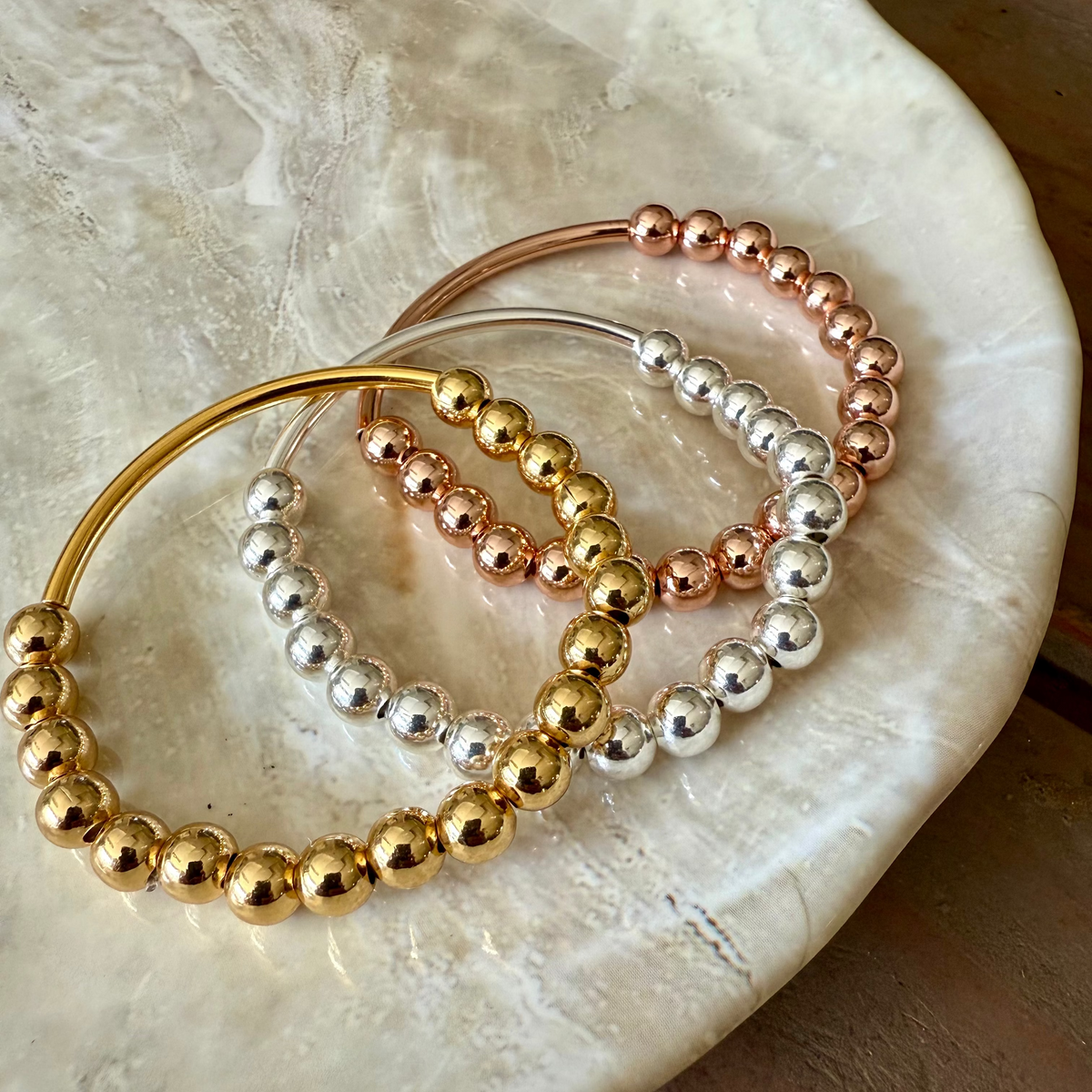 Three stretch-to-fit bracelets in gold, rose gold, and silver plated metal on a marble surface.