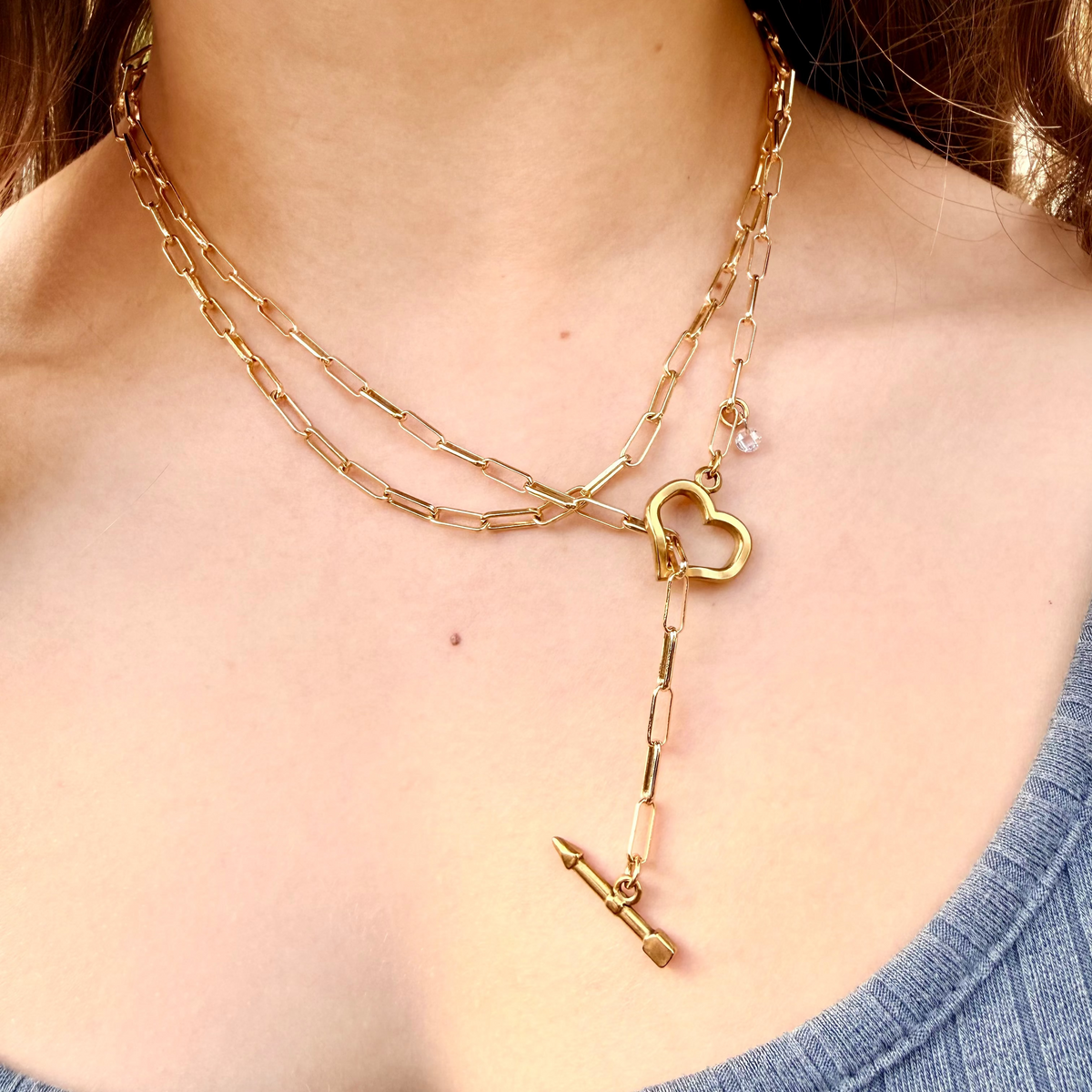 Gold chain necklace with heart and arrow pendants on a person wearing a blue top.