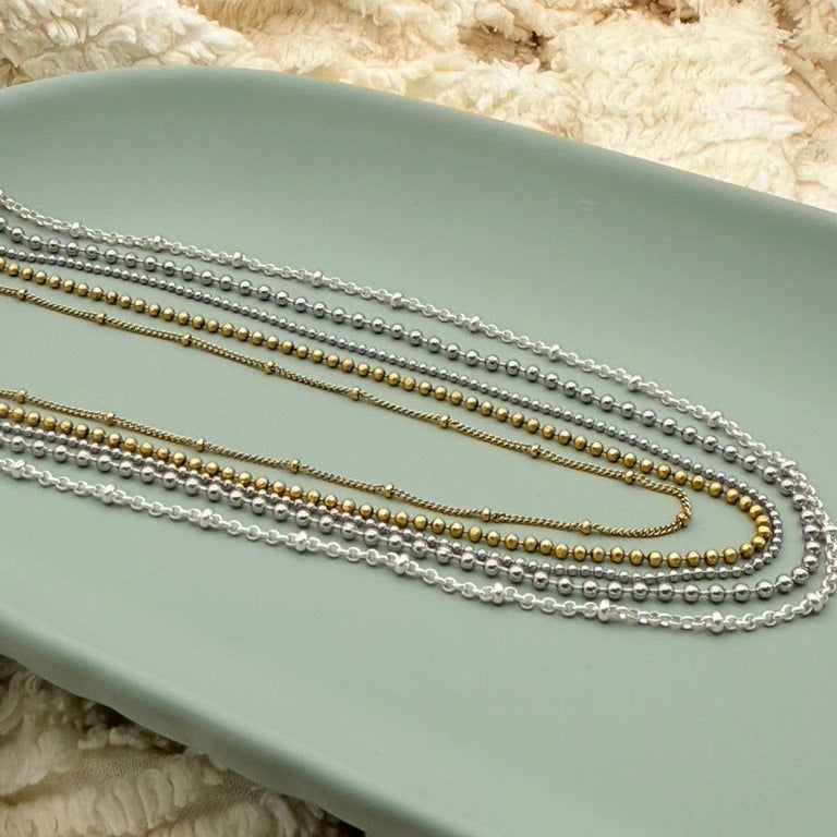 Three necklaces, two silver and one gold, on a light green surface with a textured beige background.