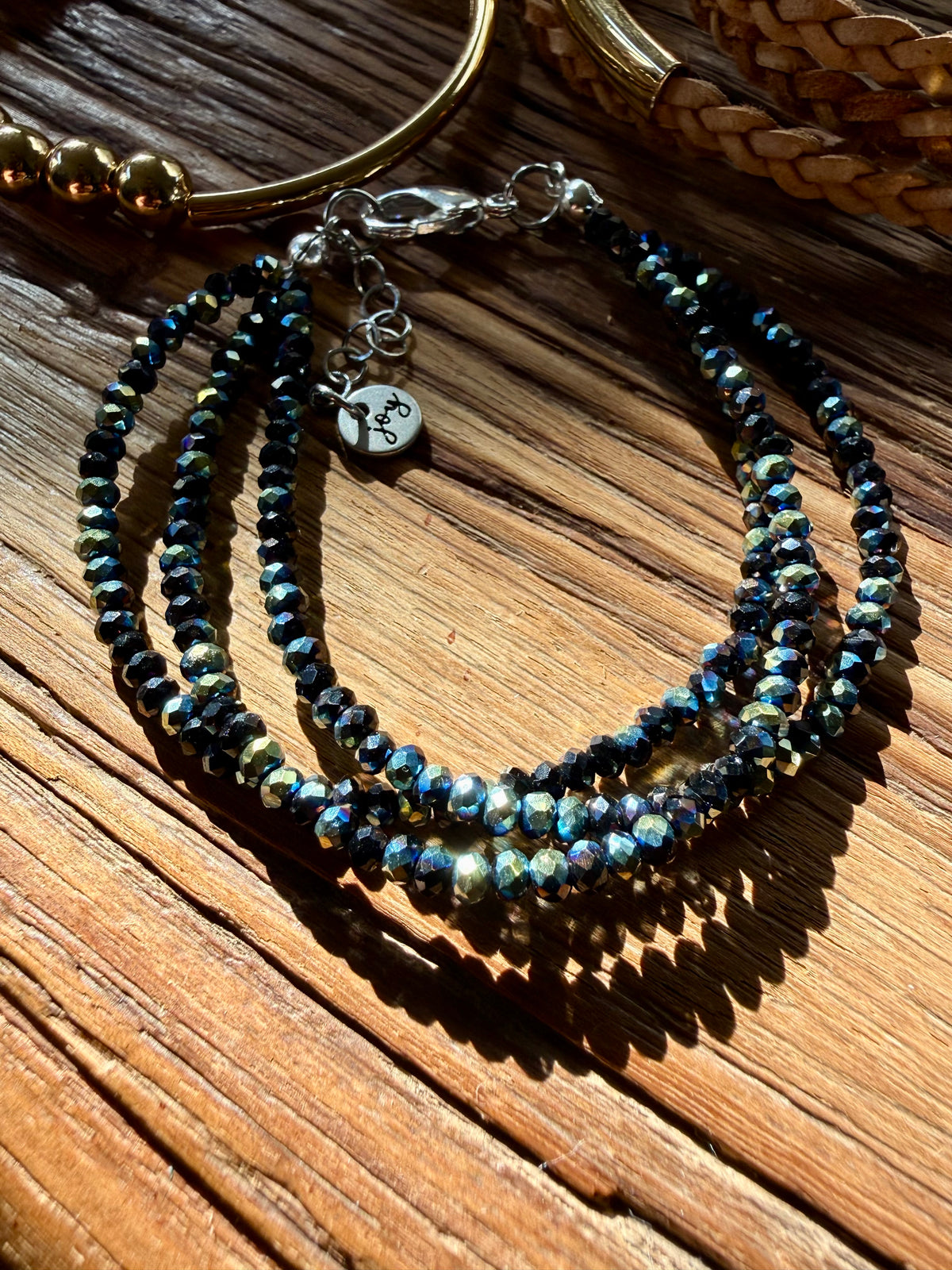 A multi-strand beaded bracelet featuring faceted dark blue and iridescent blue-green beads, accented with a silver lobster clasp and a small heart-shaped charm engraved with an anchor, displayed on a rustic wooden surface.