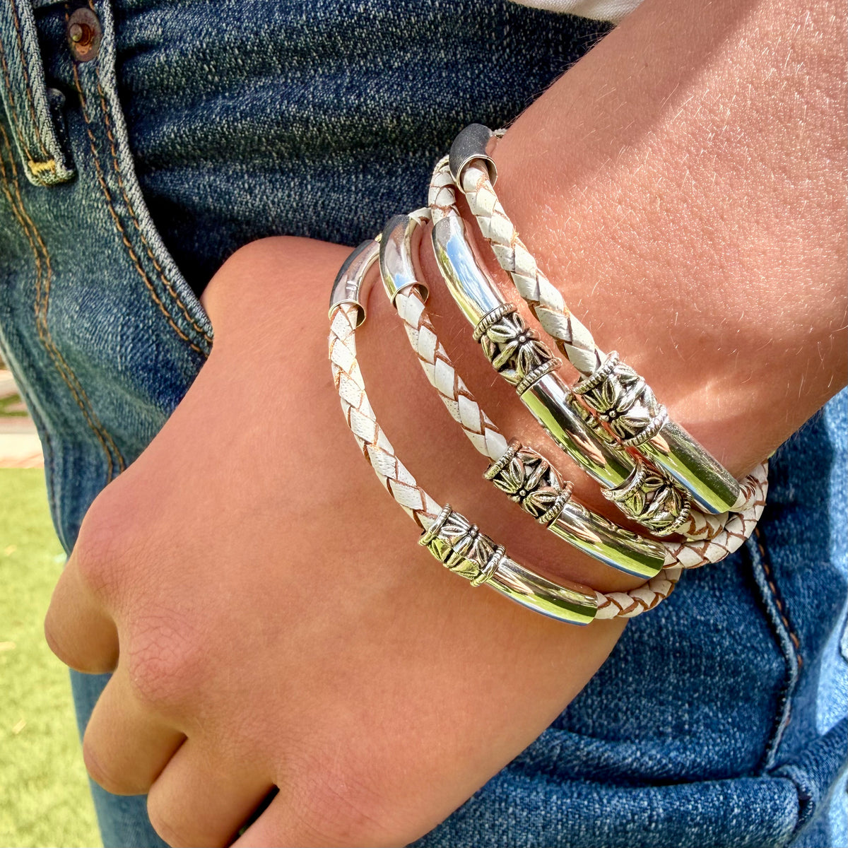 A close-up of a person's wrist wearing four bracelets: two braided white bracelets and two polished silver-tone bracelets with decorative star-like links, shown against blue denim jeans.