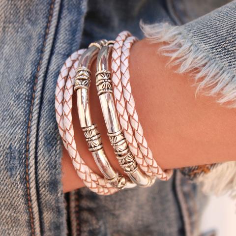 A close-up of a person's wrist wearing multiple bracelets, including two braided light pink cords and two polished silver-toned metal tubes with engraved details, styled with a frayed denim sleeve.