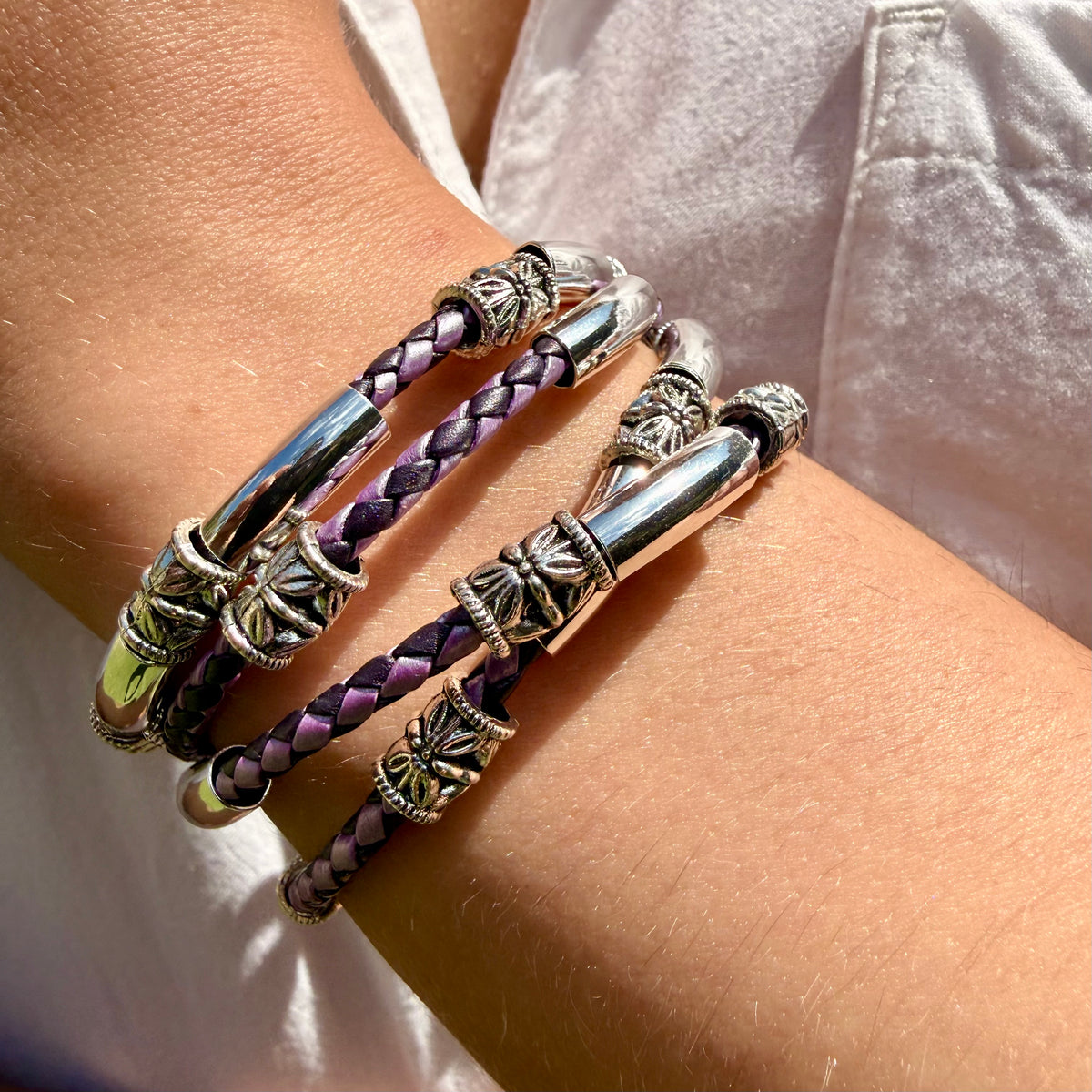 A close-up of a person's wrist wearing three braided bracelets. Two of the bracelets are purple and black braided with silver-toned metal accents, and one is a solid silver-toned bangle.