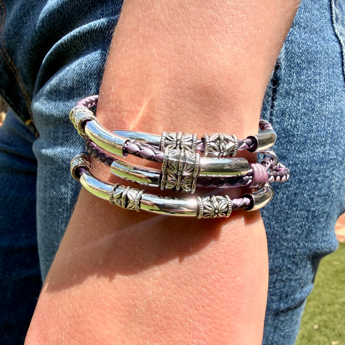 A close-up of a person's wrist wearing three stacked bracelets: two metallic silver-toned tubes with textured details and one purple braided cord bracelet with a matching bead accent.
