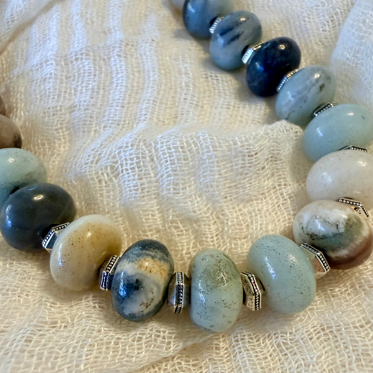 A beaded necklace featuring polished light blue, navy, cream, and marbled stone beads separated by small silver-toned metal spacers, displayed on a textured off-white fabric background.