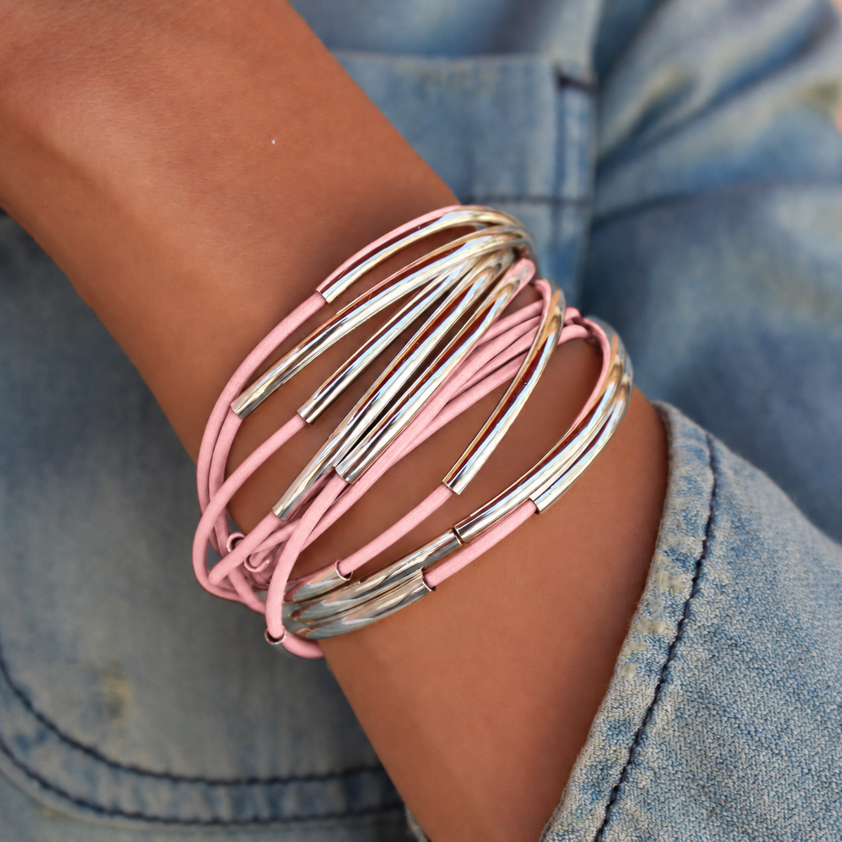 A close-up of a person's wrist wearing a stack of multi-layered bangles, featuring light pink cords and polished silver metal tubes, paired with a blue denim sleeve.