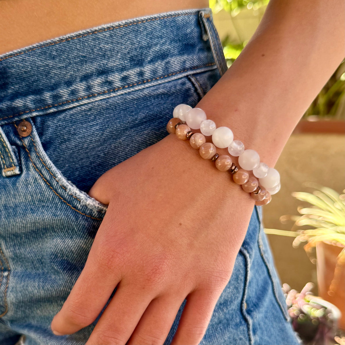 2 beaded stretch bracelets, one made of smooth, round peach-pink stone beads separated by small metallic copper color spacer beads, and the other with 2 difference sized light pink round beads shown in an outdoor background.