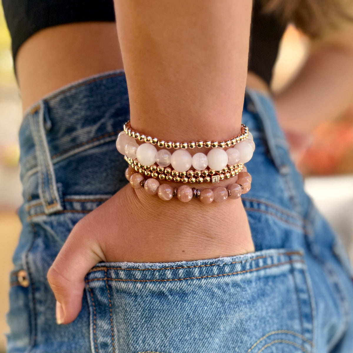 Multiple  pink gemstone and rosegold bracelets of different sizes stacked on a woman's wrist with her hand in her jean pocket. 