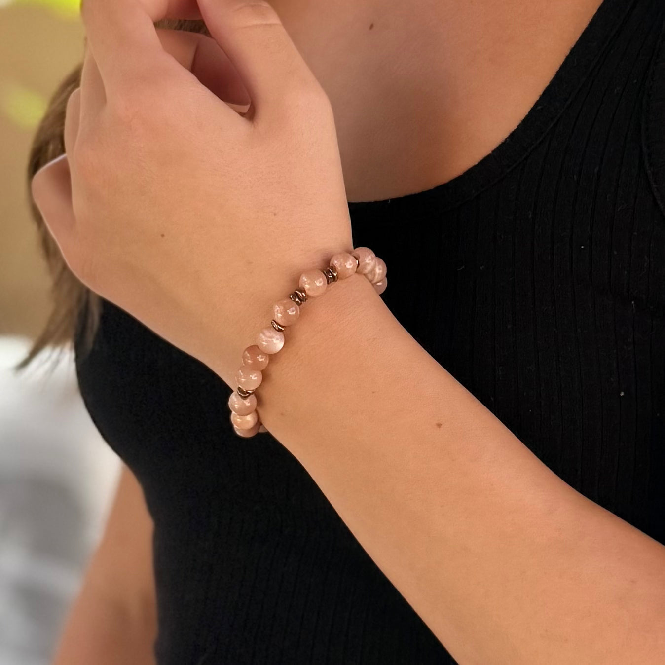 A beaded stretch bracelet made of smooth, round peach-pink stone beads separated by small metallic bronze spacer beads, shown on an outdoor background.