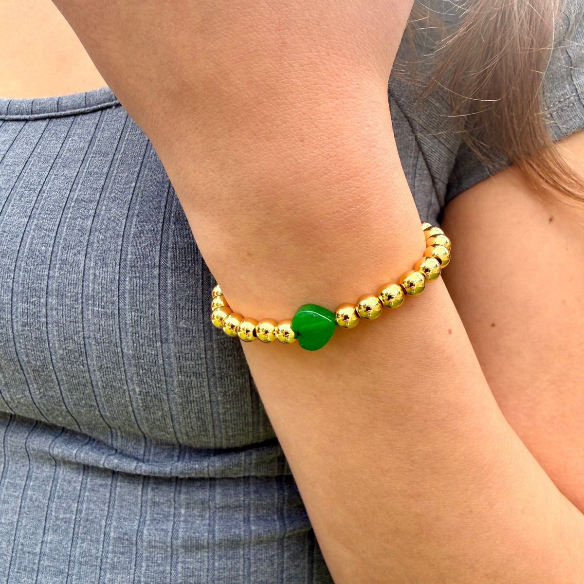 Gold beaded bracelet with a green heart charm on a wrist against a gray background