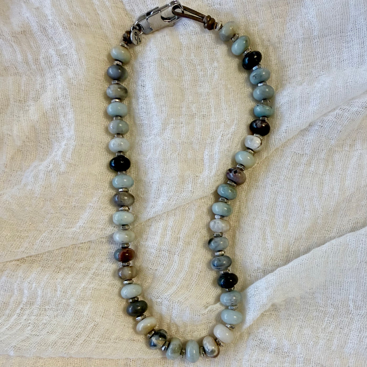 A beaded necklace made of polished round stone beads in muted gray, pale blue-green, black, and brown tones, with small metallic spacer beads and a silver-toned clasp, laid on a light beige textured fabric background.